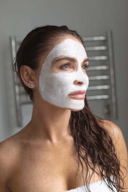 Woman with facial mask standing in bathroom, looking thoughtful. Wet hair and white towel suggest post-shower skincare routine. Ideal for use in beauty, skincare, and wellness blogs, advertisements for beauty products, and magazines promoting self-care and home spa treatments.