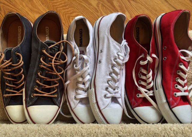 Multiple pairs of Converse sneakers in different colors arranged in a neat row along carpet and adjacent hardwood floor. This image can be used for fashion blogs, footwear advertising, casual style promotion, retail marketing, or lifestyle publications emphasizing trendy casual wear.