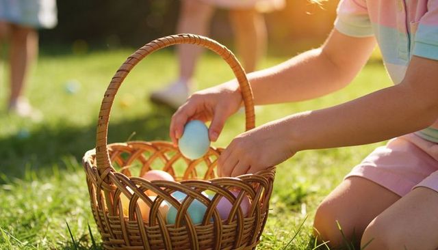 Child placing pastel easter egg into wicker basket on sunlit lawn during egg hunt
