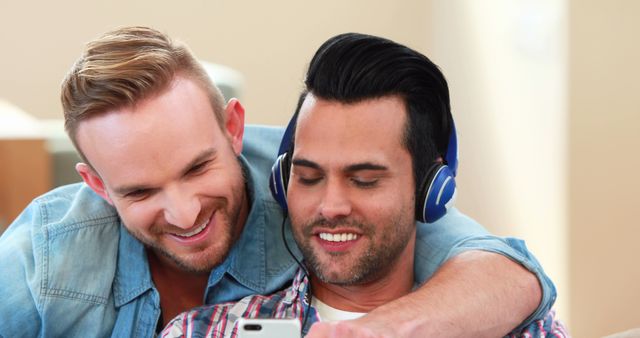 Two men are sharing earbuds and smiling while enjoying music on a mobile phone. They appear casually dressed and relaxed at home. This scene can be used in campaigns highlighting the use of technology for entertainment, promotions geared towards couples and friendships, or any visuals pertaining to modern lifestyle and connectivity.