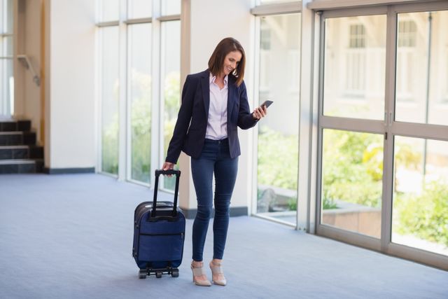 Businesswoman in professional attire carrying luggage and using mobile phone at a conference center. Ideal for themes related to business travel, corporate events, professional communication, and modern business environments. Useful for articles, blogs, and advertisements focusing on business trips, corporate mobility, and professional lifestyles.