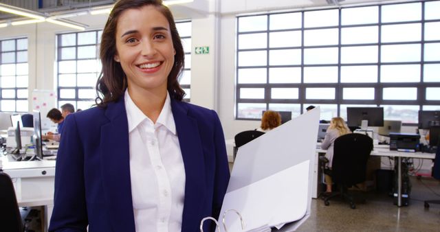 A confident businesswoman is holding documents and smiling in a modern office. This stock photo is perfect for illustrating corporate success, professional environments, or promotions. Ideal for use in business presentations, training materials, website banners, and office-related articles.