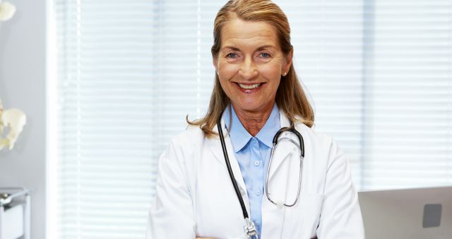 Senior female doctor smiling and wearing a white coat with a stethoscope around her neck. She appears in a medical office with blinds in the background. Suitable for articles about healthcare professionals, medical services, and health consultations.