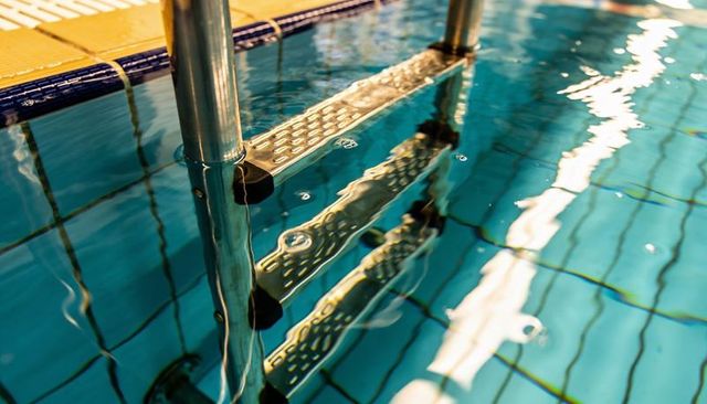 Stainless steel pool ladder reflecting in clear blue water with tile grid and bubbles