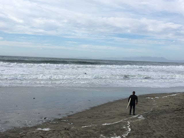 This image shows a surfer in a wetsuit standing on a sandy beach, gazing at the ocean waves under a cloudy sky. Ideal for use in content related to surfing, ocean sports, outdoor activities, beach vacations, coastal sceneries and marine weather. It can be used for travel websites, adventure sports ads, or blog posts about surfing tips and experiences.