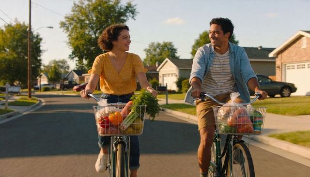 Young couple cycling with grocery-filled baskets along sunlit suburban street, smiling