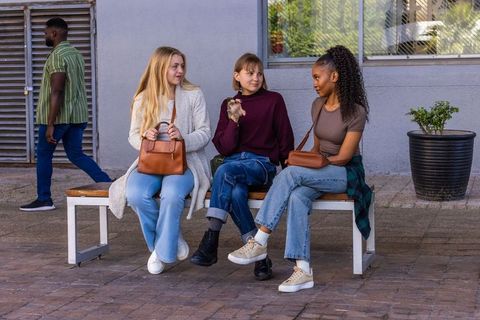 Three friends chatting on bench holding brown leather handbags on urban sidewalk