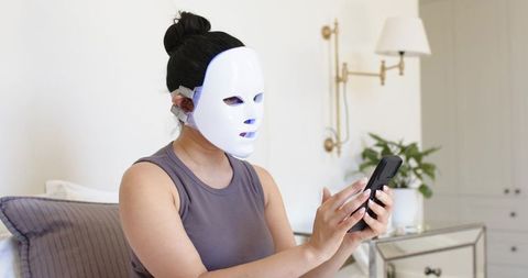 Woman wearing led facial mask using smartphone in minimalist bedroom skincare routine