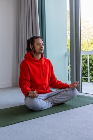 African american man meditating on yoga mat by sunlit balcony wearing red hoodie