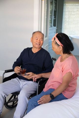 Senior asian couple sharing tea at home, man in wheelchair smiling, intimate care moment