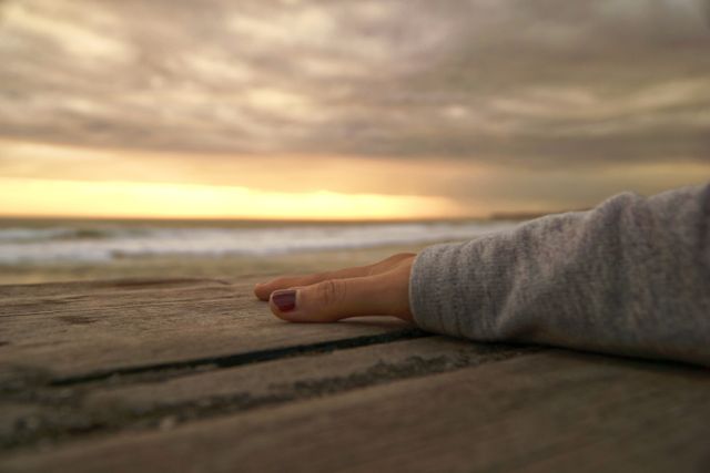A hand with painted nails gently resting on a wooden surface, with an ocean view at sunset in the background. This peaceful scene showcases a calm, tranquil moment ideal for themes of relaxation, contemplation, and nature's beauty. Useful for meditation, wellness, and travel content.