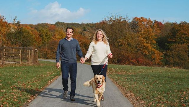 Smiling couple holding hands walking retriever on paved country path amid autumn foliage