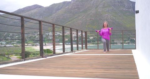 Mature woman performing lunges on coastal deck with dumbbells, ocean view fitness lifestyle