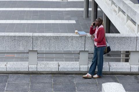 Urban commuter leaning on concrete balustrade talking on phone with coffee and notebooks