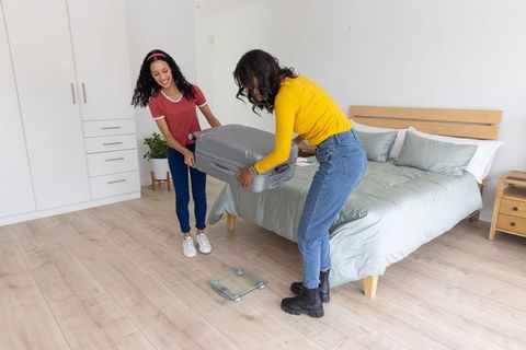Mother and teen daughter lifting suitcase onto scale in bright minimal bedroom before trip