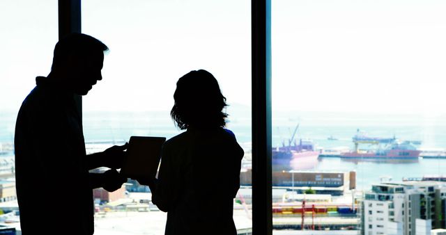 Two business colleagues are discussing a project in an office. The background shows a panoramic view of the sea and shipping port, adding a cosmopolitan and professional atmosphere. The image can be used for corporate presentations, websites highlighting teamwork and collaboration, or promotional materials for business and maritime industries.