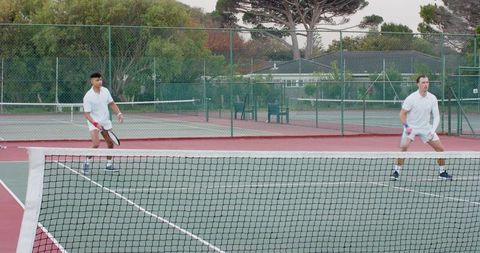 Diverse male doubles partners standing ready on outdoor hard court holding rackets concentrating
