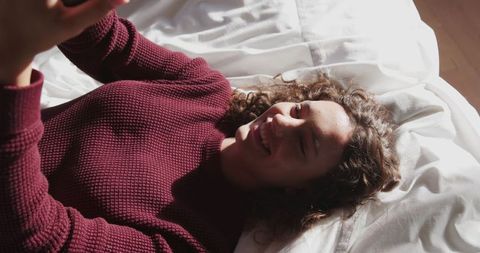 Woman Enjoying Relaxed Moment on Bed with Smartphone in Sunlight