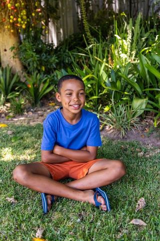 African american boy sitting cross-legged on lawn smiling in backyard garden