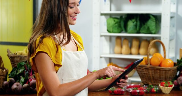 Woman wearing apron smiling while using tablet in organic store displaying fresh vegetables and fruits. Ideal for themes related to small business operations, technology in retail, organic farming, healthy eating, and market management.