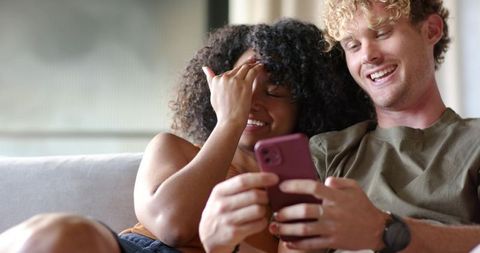 Laughing diverse couple sharing smartphone on sofa, cozy candid home moment