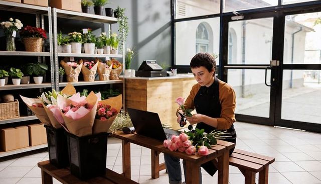 Florist arranging pink rose bouquet while managing online orders on laptop in sunny shop