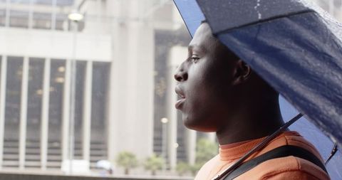 African American man standing under blue umbrella, gazing across rainy downtown in orange sweater