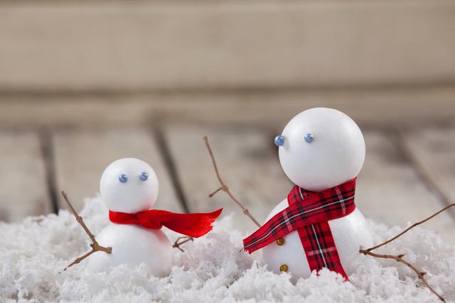 This image shows two snowmen with scarves sitting in fake snow. The snowmen are adorned with red and plaid scarves, and have small twig arms. This image can be used for holiday cards, winter-themed decorations, Christmas promotions, or festive social media posts.