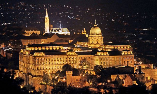Royal Palace in Budapest glowing with lights at night, surrounded by cityscape. Ideal for travel brochures, websites promoting tourism, background images for European cities, educational materials on historic landmarks, and digital postcards.