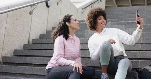 African American Women Sitting on City Stairs Taking Selfie in Hoodies and Activewear