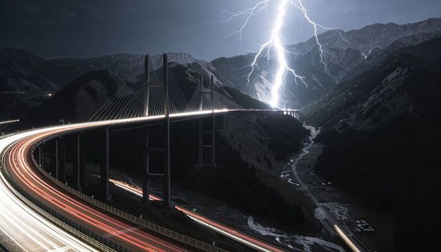 Curving cable-stayed viaduct carrying light trails over mountain gorge lightning strike at night