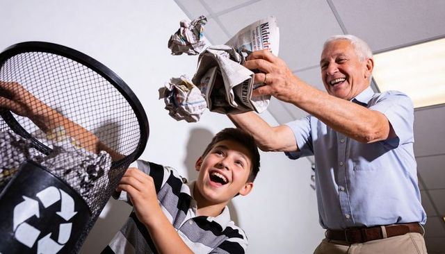 Senior man mentoring teen tossing crumpled newspaper into recycling bin, smiling