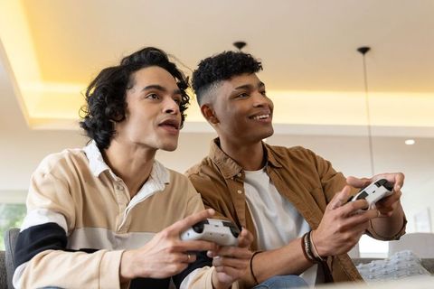 Two friends playing video games on sofa with white controllers, laughing and competing