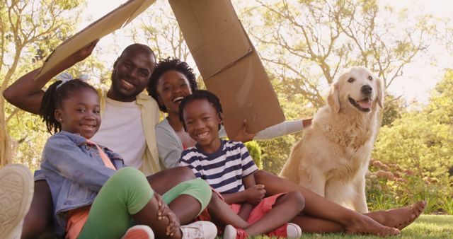 A happy African American family enjoys a sunny day outdoors under a makeshift cardboard fort, with copy space. Smiles abound as the children, parents, and their golden retriever create memories in their backyard.