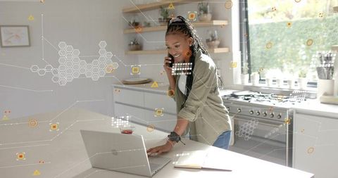 Young woman multitasking on laptop and phone in sunlit modern kitchen home office remote work