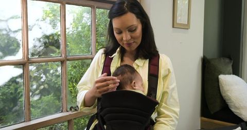 Asian mother comforting infant in baby carrier at bright home window
