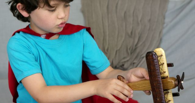 Young boy wearing red cape, engaging in creative play with wooden toy airplane. Perfect for use in themes related to childhood imagination, creativity, and adventure. Suitable for parenting blogs, educational materials, and advertisements for toys or children's activities.