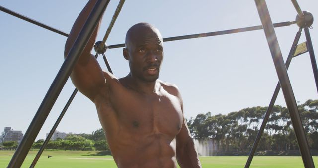 Fit shirtless african american man exercising on climbing frame outdoors, taking a rest. cross training for fitness at a sports field.