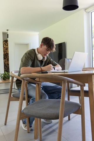 Teen studying at home with laptop and headphones at sunlit wooden table
