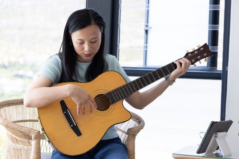 Mature asian woman practicing acoustic guitar by window with tablet for online lesson