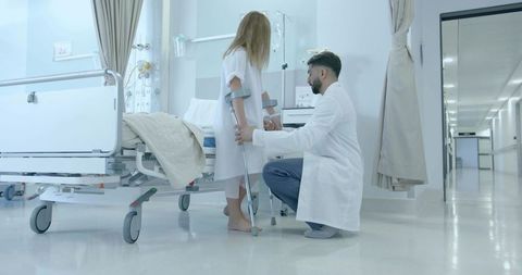 Kneeling Doctor Helping Young Patient Using Crutches in Hospital Ward with IV Stand
