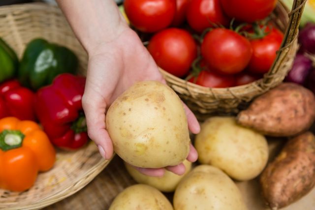 Close-up of a hand holding a fresh potato at a vegetable stall, surrounded by various colorful vegetables like tomatoes, bell peppers, and sweet potatoes. Ideal for use in articles or advertisements about healthy eating, farmers markets, organic produce, and local food sourcing.