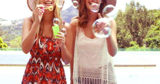 Two young women are enjoying a sunny day outdoors, blowing bubbles. They are dressed in casual summer attire and wearing hats. This can be used for promoting summer activities, friendship, leisure, and carefree lifestyles. Ideal for travel, fashion, and lifestyle blogs or magazines.