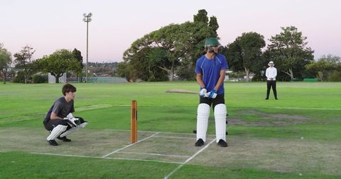 Evening cricket batsman batting at crease with wicketkeeper and umpire on green field