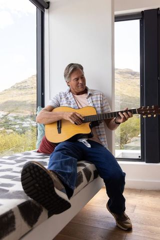 Mature man playing acoustic guitar on window bench in modern home with mountain view