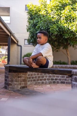 African american boy sitting barefoot on brick bench in sunny courtyard, minimalist lifestyle