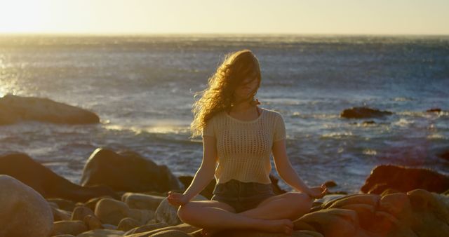 Young Caucasian woman meditates on a rocky beach at sunset. She finds tranquility in a serene outdoor setting by the sea.