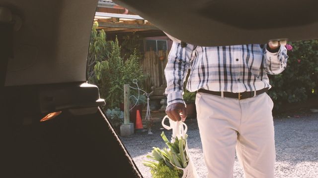 A senior man is packing grocery bags into a car trunk, captured from a low angle. He is dressed casually in a plaid shirt and beige trousers, indicating a relaxed, everyday activity. The scene represents convenience and everyday errands, emphasizing themes around organic food shopping, self-reliance, and active lifestyle. This image can be used for advertising shopping convenience, promoting local businesses, or blogs discussing day-to-day activities and healthy lifestyle choices.