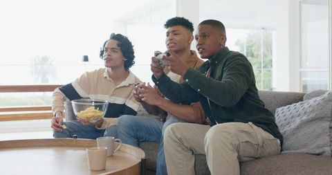 Diverse friends playing multiplayer game on couch, sharing snacks in sunlit living room