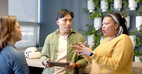 Diverse team collaborating in modern workspace, woman presenting and gesturing with tablet